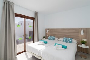Une chambre d’hôtel avec deux lits, une tête de lit en bois, une petite table de chevet avec une lampe, et une vue à travers une porte vitrée.