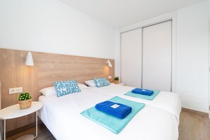 Ein Hotelzimmer mit einem hölzernen Kopfteil, zwei Betten mit weißer Bettwäsche und gemusterten Kissen, einem kleinen Nachttisch mit einer Lampe, einer Steckdose und einer Topfpflanze.
