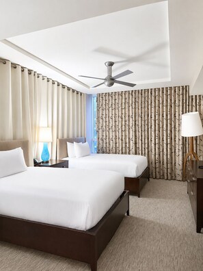 Ein Hotelzimmer mit einem großen Bett, einem kleineren Bett, einem Deckenventilator, einem Schreibtisch mit Lampe und Blick nach außen.