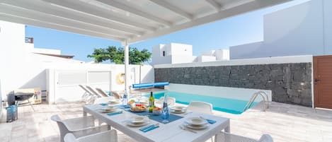 A covered outdoor dining area with a pool, white furniture, and a stone wall.