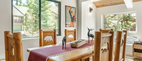 Un comedor con sillas de madera y un banco, un mantel rojo y vistas a un bosque a través de amplios ventanales.