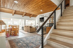 A spacious living area with a wooden ceiling, a staircase with a black railing, and a view of the outside through large windows. - Newly Remodeled Walk to Ski Condo 615 (Mammoth Lakes)