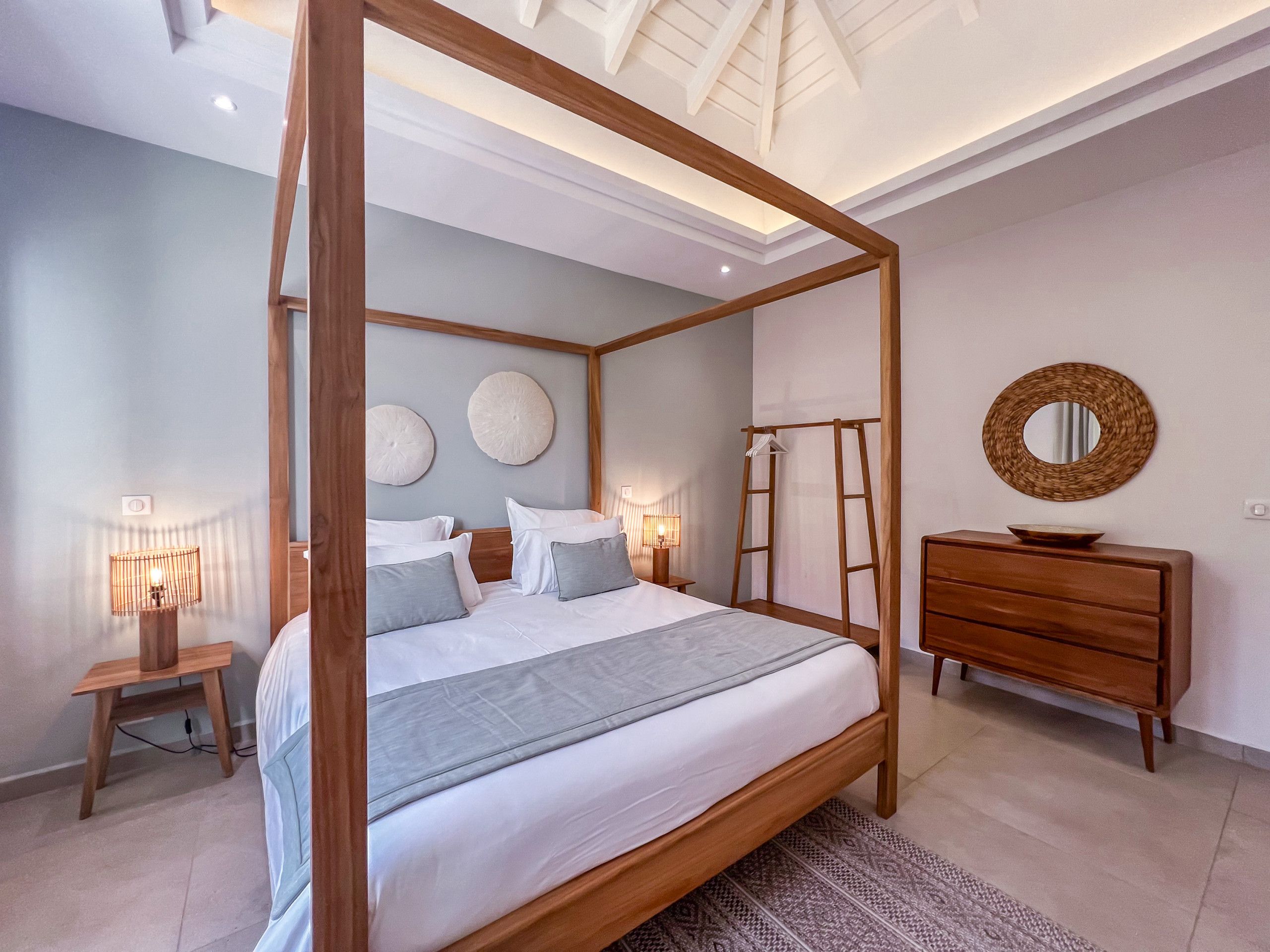 Una cama con dosel con estructura de madera, una mesita de noche con lámpara, un aparador y un espejo redondo en la pared.