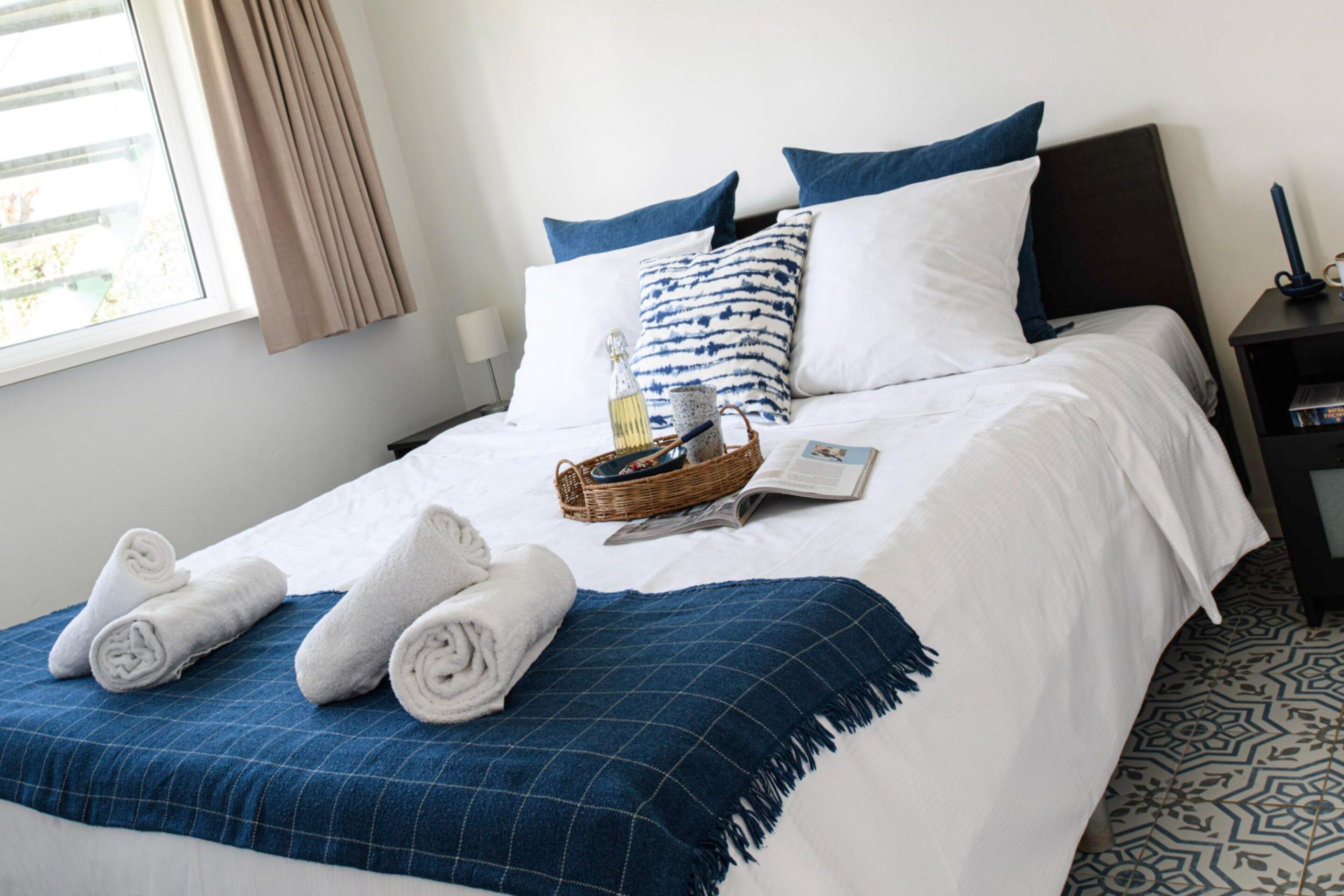 A neatly made bed with white bedding, a blue checkered throw, and rolled towels.