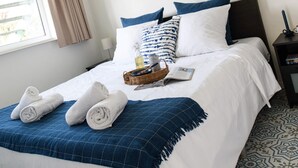Cama bem arrumada com roupa de cama branca, manta xadrez azul e toalhas enroladas.