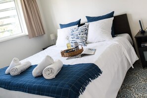 Una cama bien hecha con ropa blanca, un plaid azul de cuadros y toallas enrolladas.