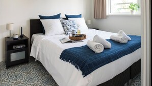 Cama bem arrumada com manta azul, travesseiros e toalhas dobradas. Há uma mesa de cabeceira com abajur, livro e um cesto com uma garrafa. O quarto tem piso com ladrilhos estampados e uma janela com cortinas.
