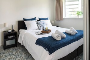 Una cama bien hecha con una manta azul, almohadas y toallas enrolladas. Hay una mesita de noche con una lámpara, un libro y una cesta con una botella. La habitación tiene baldosas de piso estampadas y una ventana con cortinas.