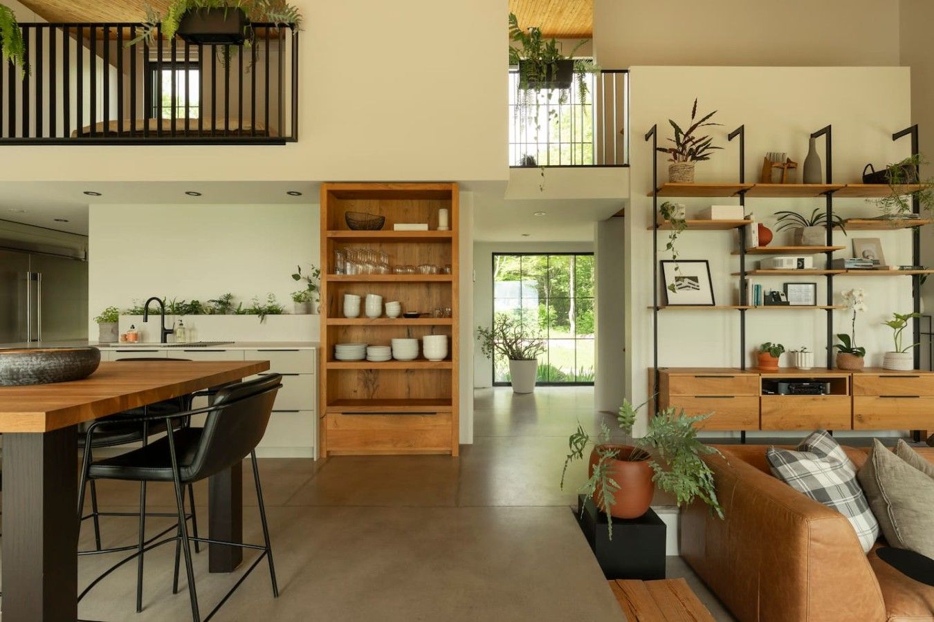 A modern interior with a kitchen, dining area, and living space. There are wooden shelves with plants and decorative items, a sofa with cushions, and a dining table with chairs.