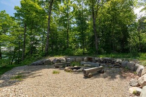 A forested campsite with a gravel ground, wooden benches, and a fire pit. - TOP LUXE mountain + lake VIEW | Hot Tub • Fire pit (Les Laurentides Regional County Municipality)