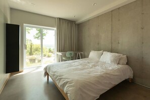 A bedroom with a large bed, a chair, a TV, and a view through a glass door.