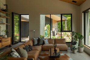 A modern living room with a large sectional sofa, a coffee table, and a view of a garden through large windows. - TOP LUXE mountain + lake VIEW | Hot Tub • Fire pit (Les Laurentides Regional County Municipality)