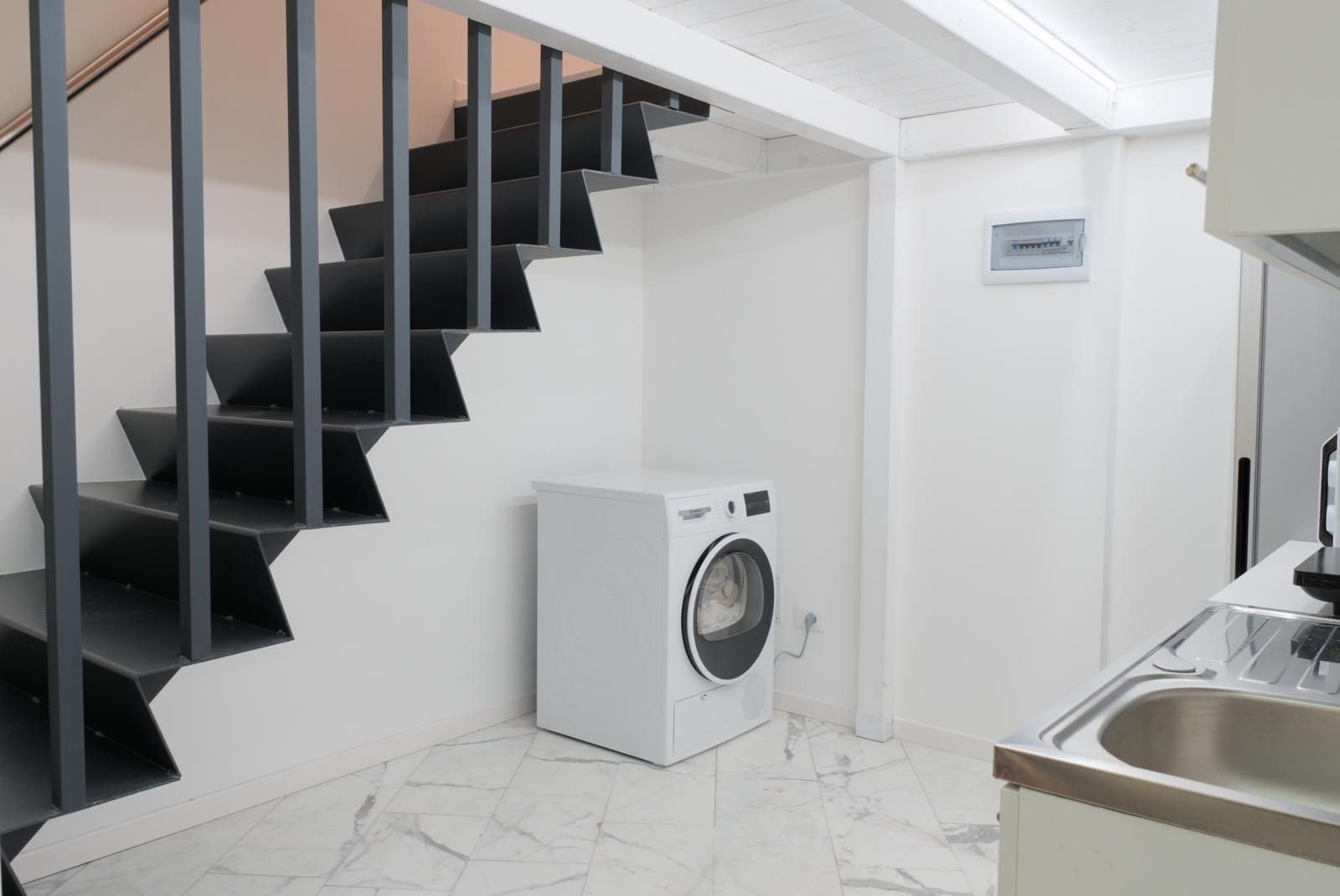 A modern kitchen with a built-in washing machine, a stainless steel sink, and a staircase with black railings.