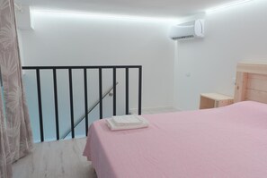 Quarto com colcha rosa, cabeceira de madeira, mesa de cabeceira pequena e janela com cortina.