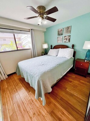 A bedroom with a bed, a ceiling fan, a nightstand with a lamp, and a window with curtains. - 2 bedroom condo in the heart of downtown Kailua Kona (Kailua Kona)