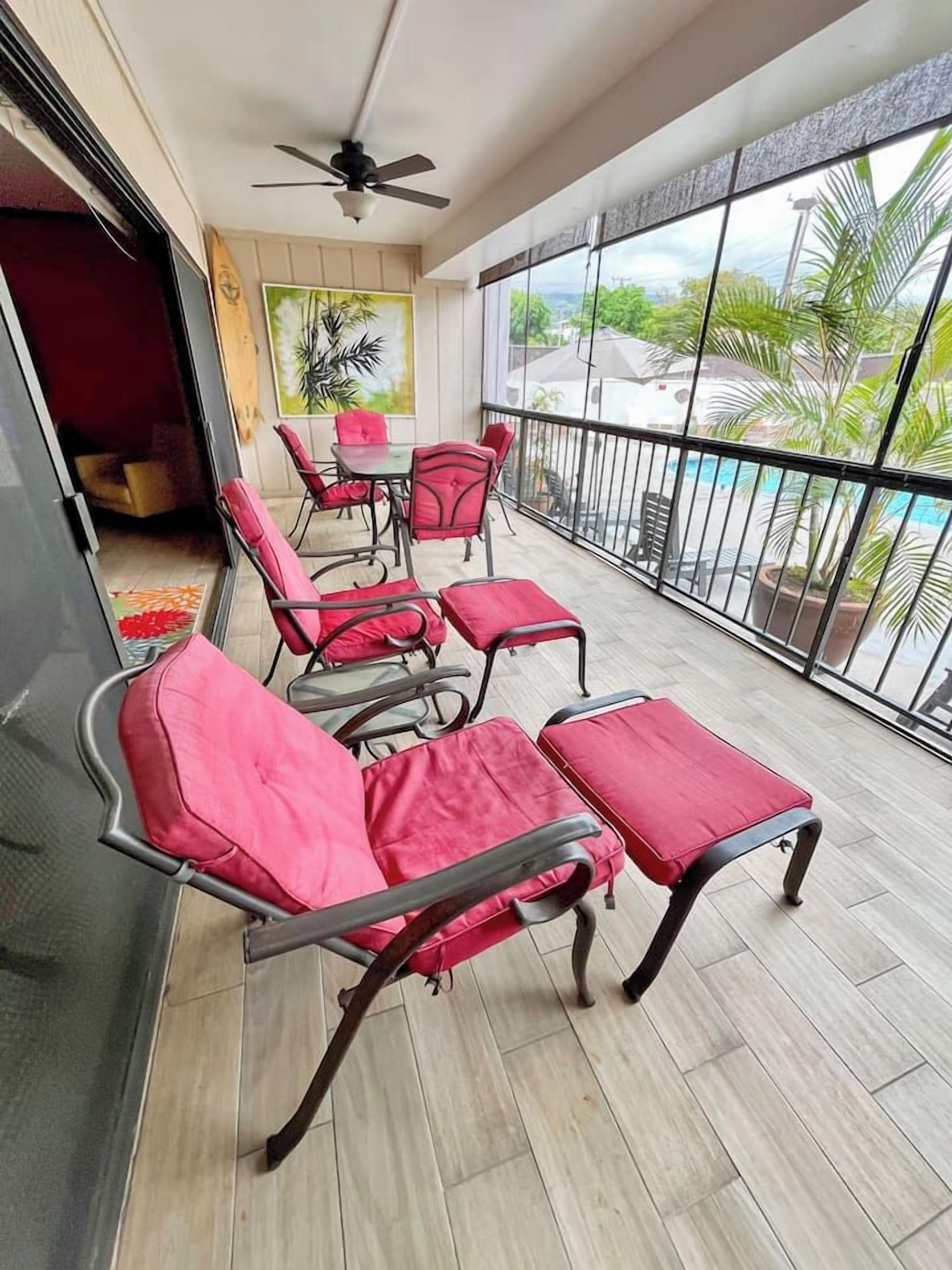 A balcony with red cushioned chairs and a table, a ceiling fan, and a view of a swimming pool and greenery.