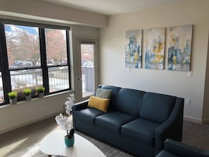 A living room with a dark blue sofa, a white coffee table, and a window with a view of parked cars and trees. - Modern Apt a Block from St. Pat's Hospital #202 (Missoula)