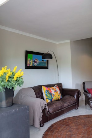 A living room with a brown leather sofa, a colorful throw pillow, a black floor lamp, a framed painting on the wall, and a potted plant with yellow flowers.