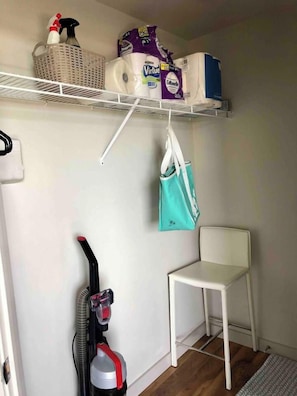A small room with a white chair, a vacuum cleaner, a shelf with cleaning supplies, and a hanging bag. - Modern Apt a Block from St. Pat's Hospital #302 (Missoula)
