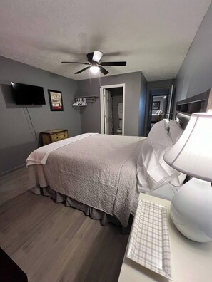 Een slaapkamer met een bed, een bureau met een lamp, een plafondventilator, een televisie en een deur naar een badkamer.