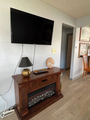 Un salon avec un téléviseur à écran plat fixé au mur, un meuble en bois orné d’une assiette décorative et une lampe posée sur le dessus.