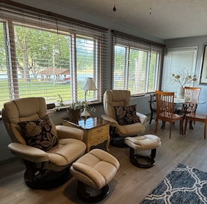 Un salon comprenant deux fauteuils beiges, un petit pouf, une table basse en bois et une table à manger avec des chaises.