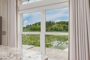 A room with a large window overlooking a pond with lily pads and a forested area. - The Waterline | lakefront, dock & hot tub, pets ok (Lincoln City)
