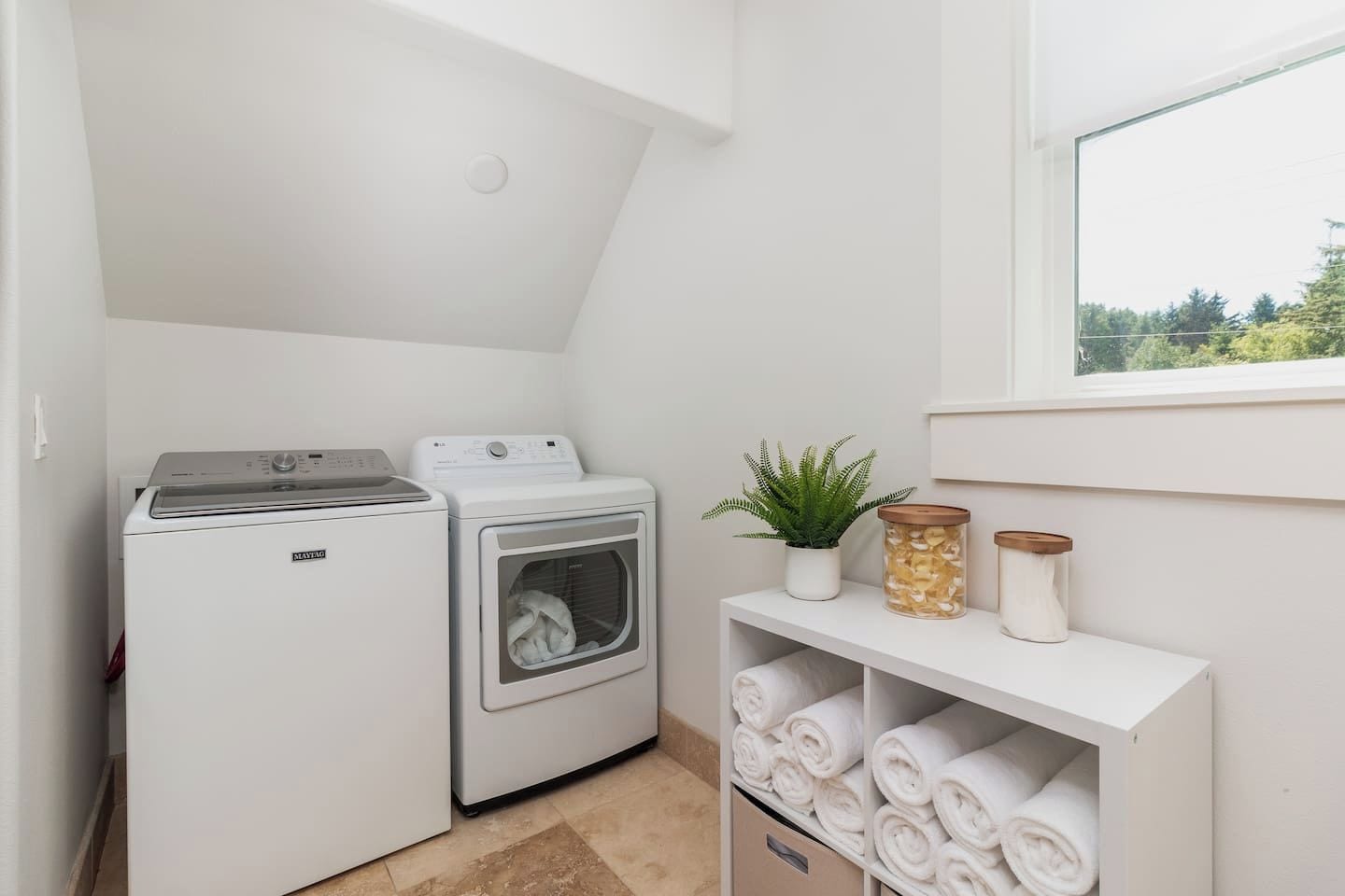 A laundry room with a front-loading washer and dryer, a shelf with folded towels, a potted plant, and a window with a view of trees.