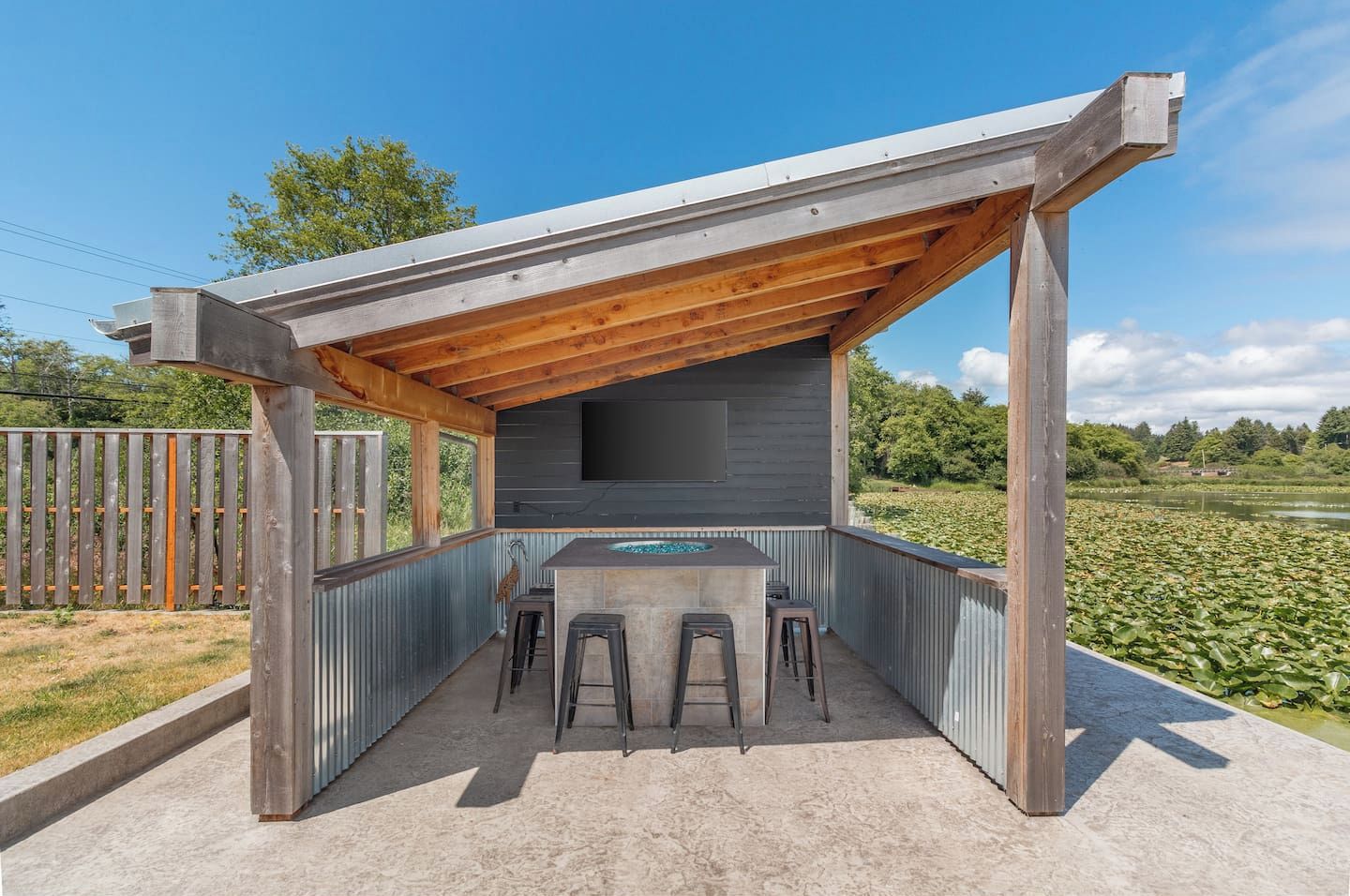 Una cocina exterior techada con encimera de concreto, taburetes de bar y un televisor montado en la pared.