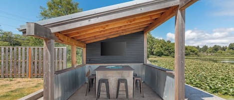Eine überdachte Außenküche mit Betonarbeitsplatte, Barhocker und einem an der Wand montierten Fernseher.