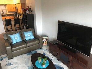 Een woonkamer met een bank, een salontafel met planten, een televisie op een kast, en een keuken op de achtergrond.