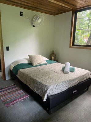 A bedroom with a bed, a bedside table, a lamp, a window, and a ceiling fan. - Cabin In The Wata (Punta Cocles)