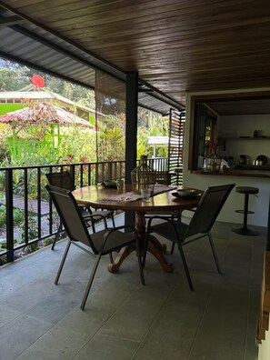Un espace extérieur abrité, aménagé pour les repas, avec une table et des chaises, une vue sur la verdure et un coin cuisine en arrière-plan.