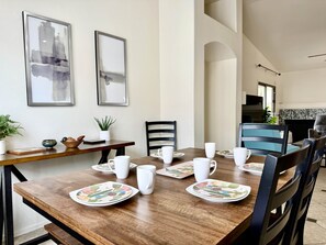 A dining area with a wooden table set for four, white plates with floral designs, and white mugs. There are two framed abstract paintings on the wall, a potted plant, and a small decorative bowl on the side table. - Dove Mountain #12361 (Marana)