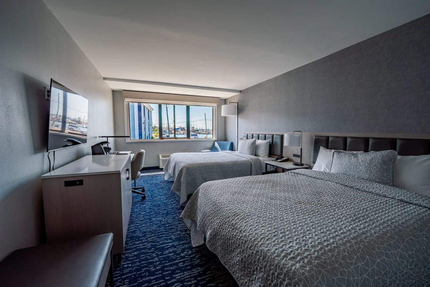 Quarto de hotel com duas camas, uma escrivaninha, uma cadeira, uma televisão e uma janela com vista para a cidade.
