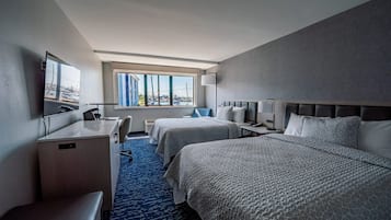 Quarto de hotel com duas camas, uma escrivaninha, uma cadeira, uma televisão e uma janela com vista para a cidade.