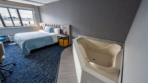 A hotel room with a bed, a desk, a chair, a large bathtub, and a view of the marina. - Harbor View - King Bed With Jacuzzi (Fiarhaven)