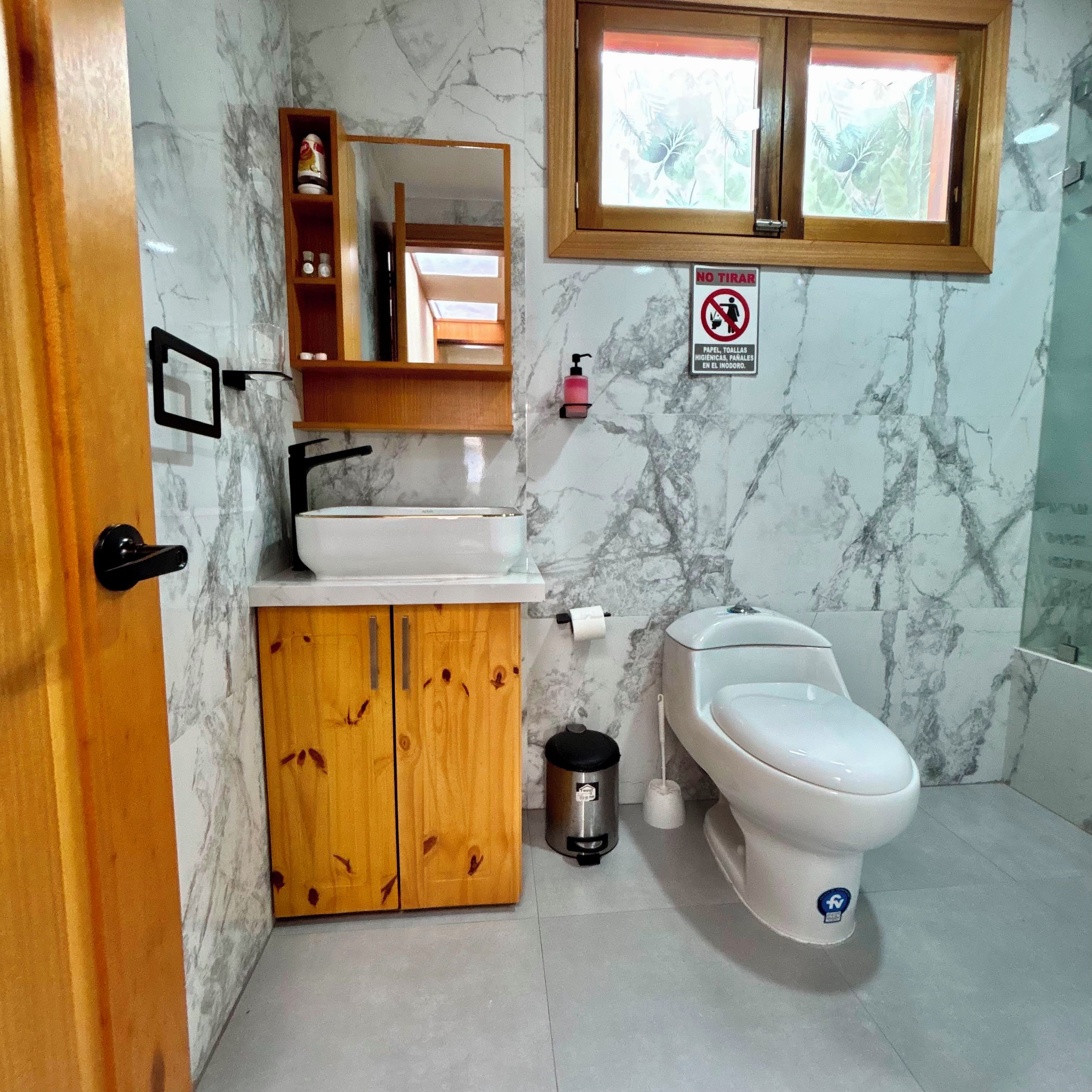 A bathroom with a marble wall, a wooden cabinet, a sink, a toilet, and a window with a view of greenery.
