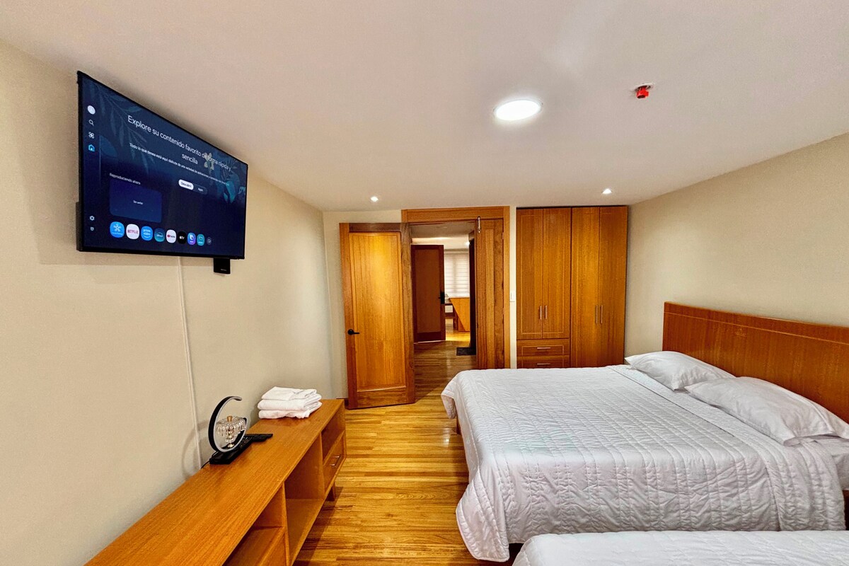 A hotel room with two beds, a TV mounted on the wall, a wooden bench, and a wooden wardrobe.