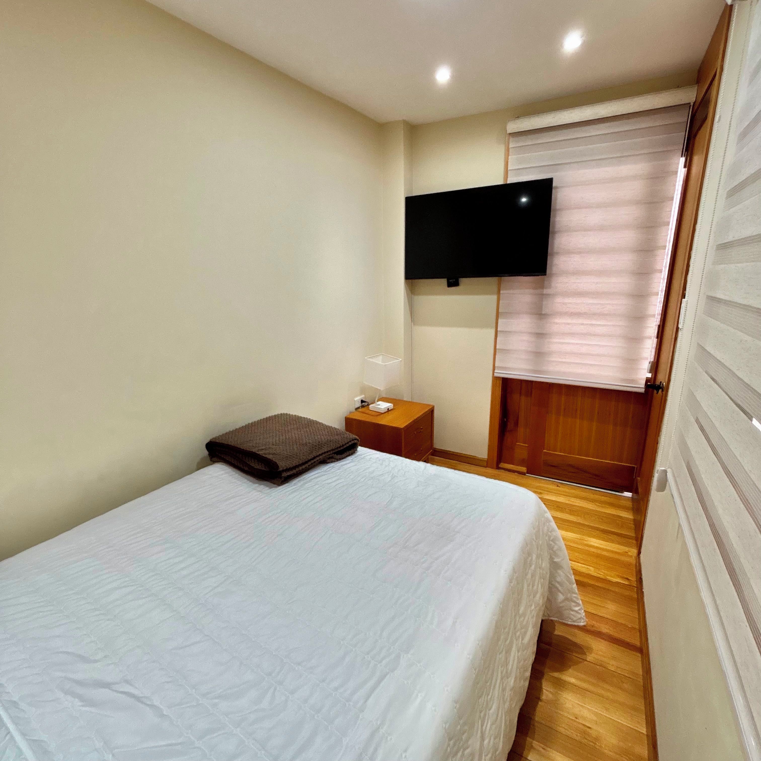 Un dormitorio con cama, mesita de noche, televisor montado en la pared y ventana con persianas.