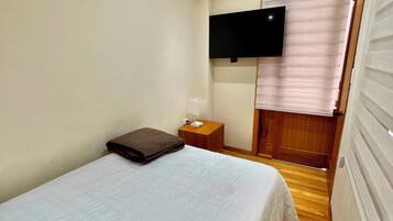 Un dormitorio con cama, mesita de noche, televisor montado en la pared y ventana con persianas.