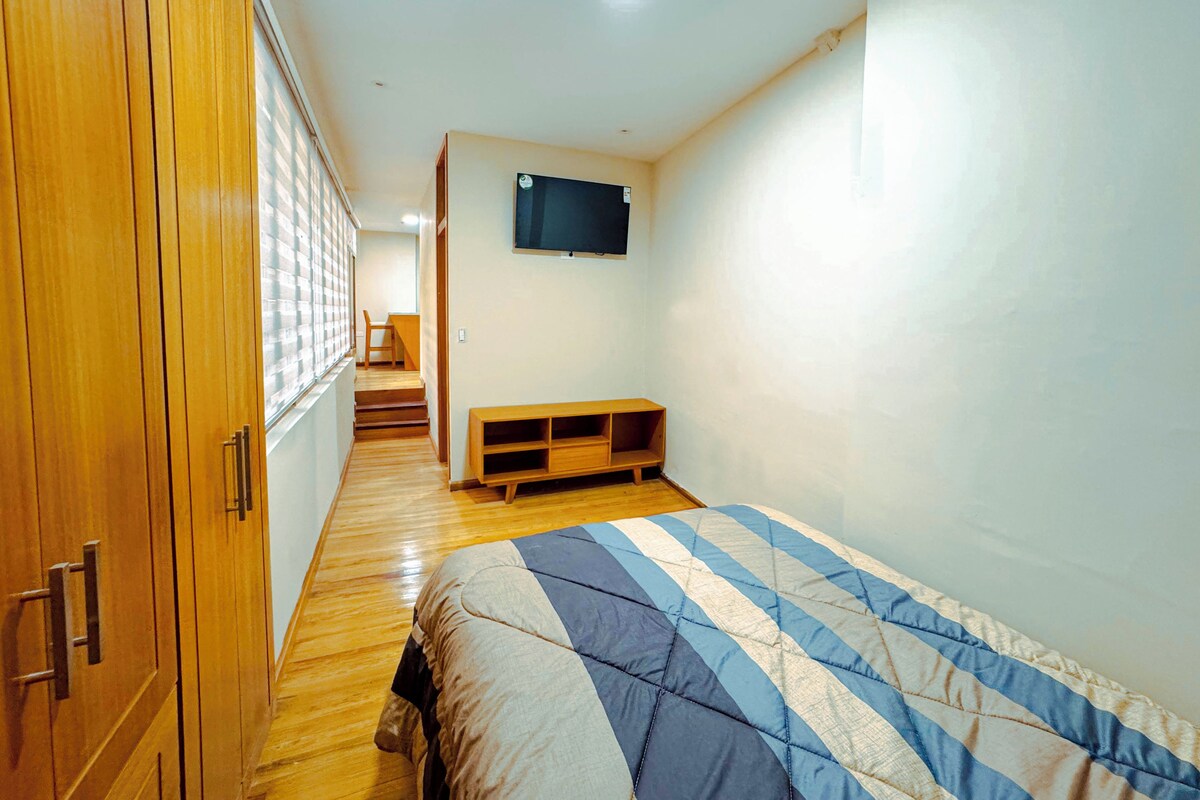 A bedroom with a bed, wooden wardrobe, a television on the wall, and a staircase leading to another room.