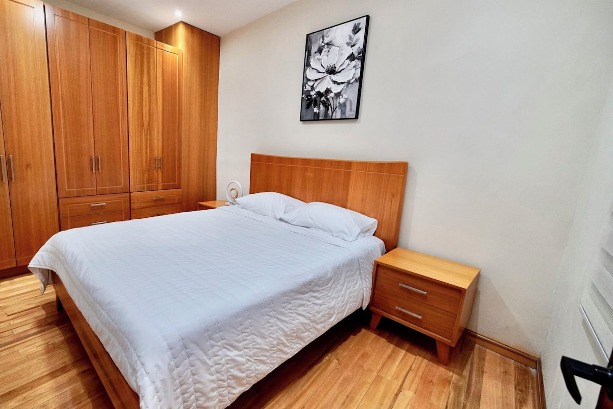 A bedroom with a wooden wardrobe, a bed with white bedding, a bedside table, and a wall-mounted artwork.