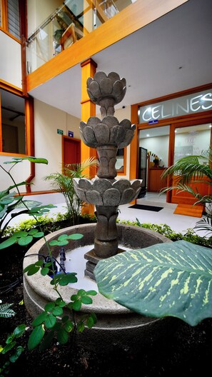 Ein springender Steinbrunnen mit mehrstufigem Design, umgeben von Topfpflanzen, in einem Eingangsbereich mit einer Rezeption und einem Schild mit der Aufschrift 'CELINES'.