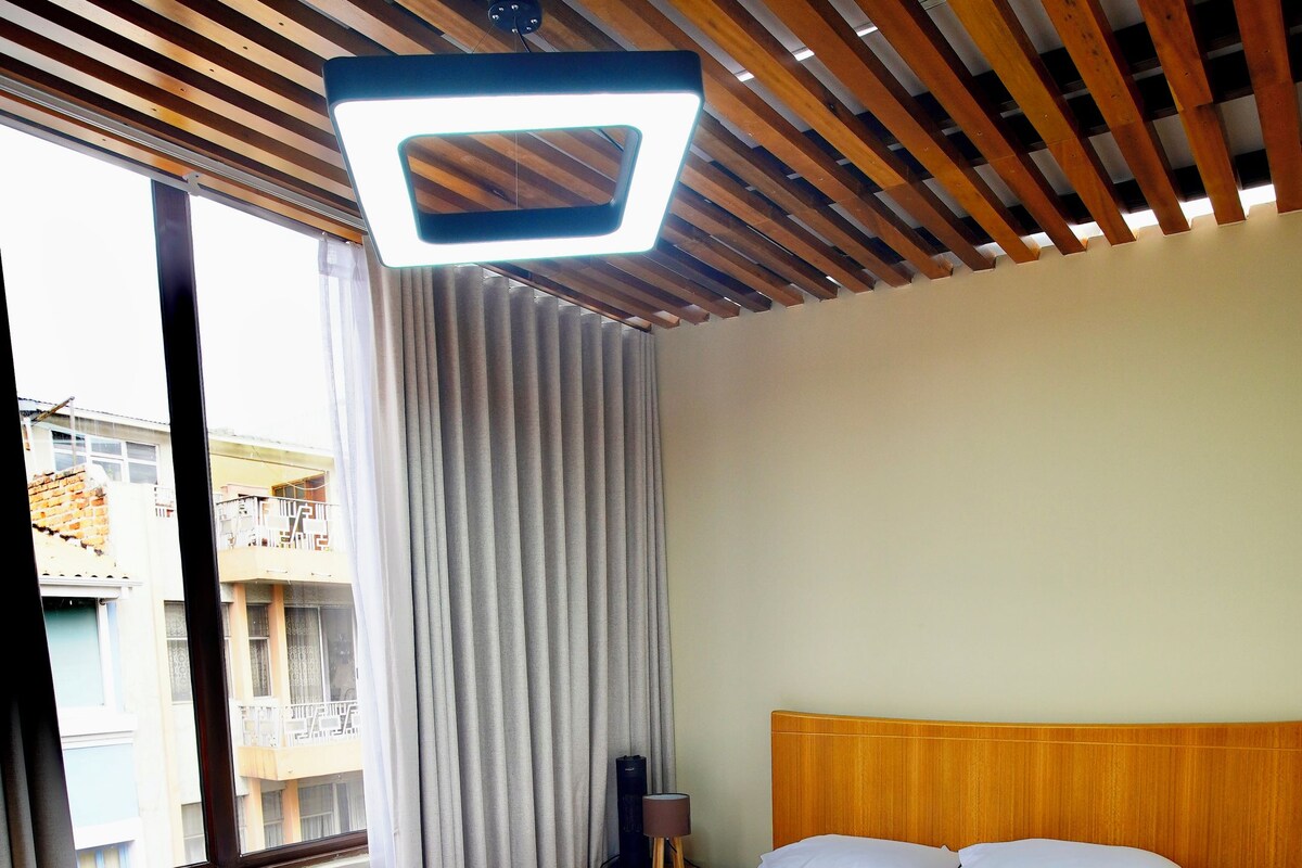 A hotel room with a wooden ceiling, a bed with white bedding, a nightstand with a lamp, and a view of buildings through a window.