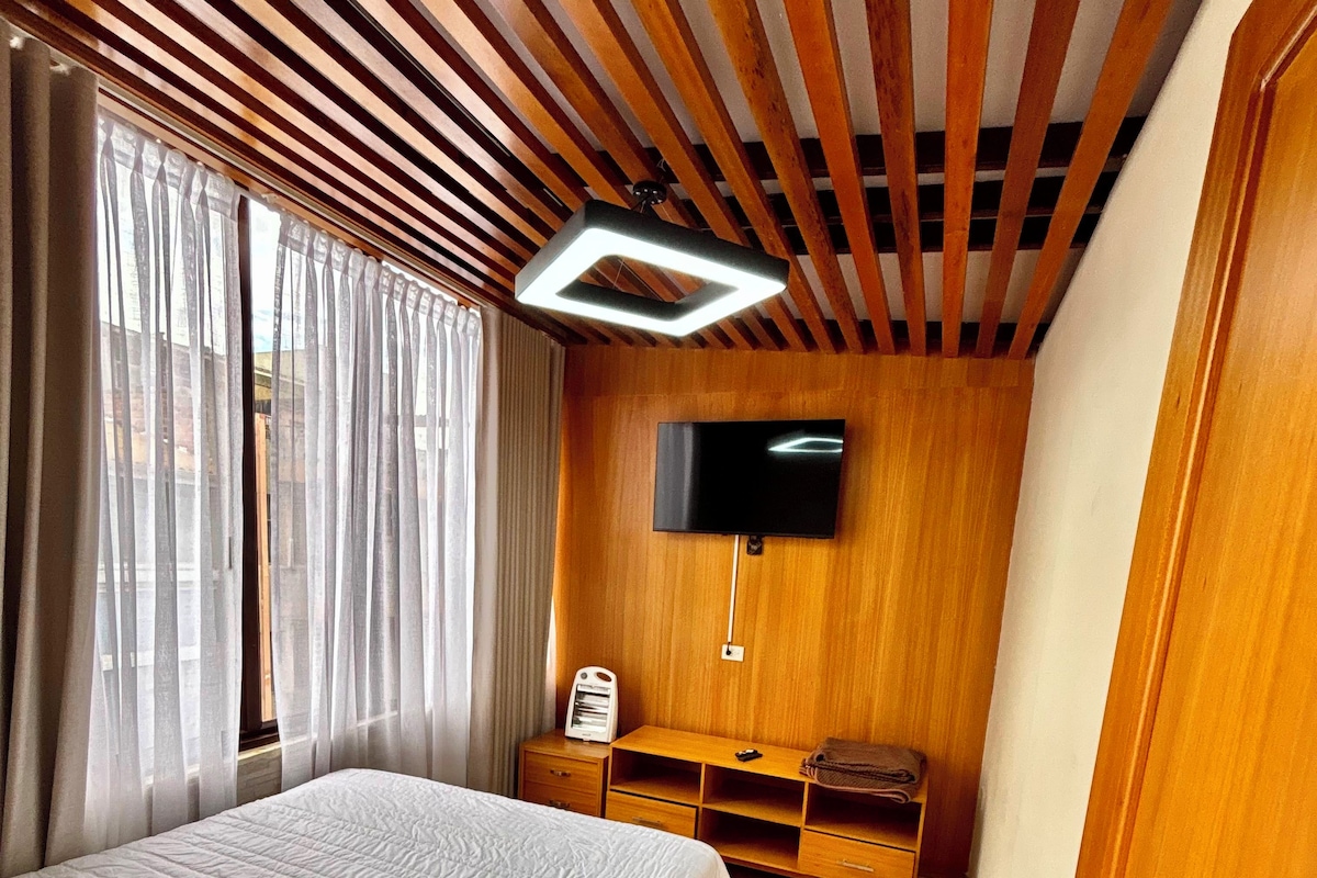 A hotel room with a bed, a TV mounted on the wall, a wooden wardrobe, and a bedside table.