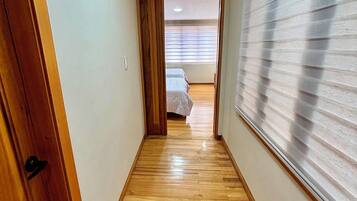 Un pasillo con suelo de madera que lleva a un dormitorio con cama y ventana con persianas.