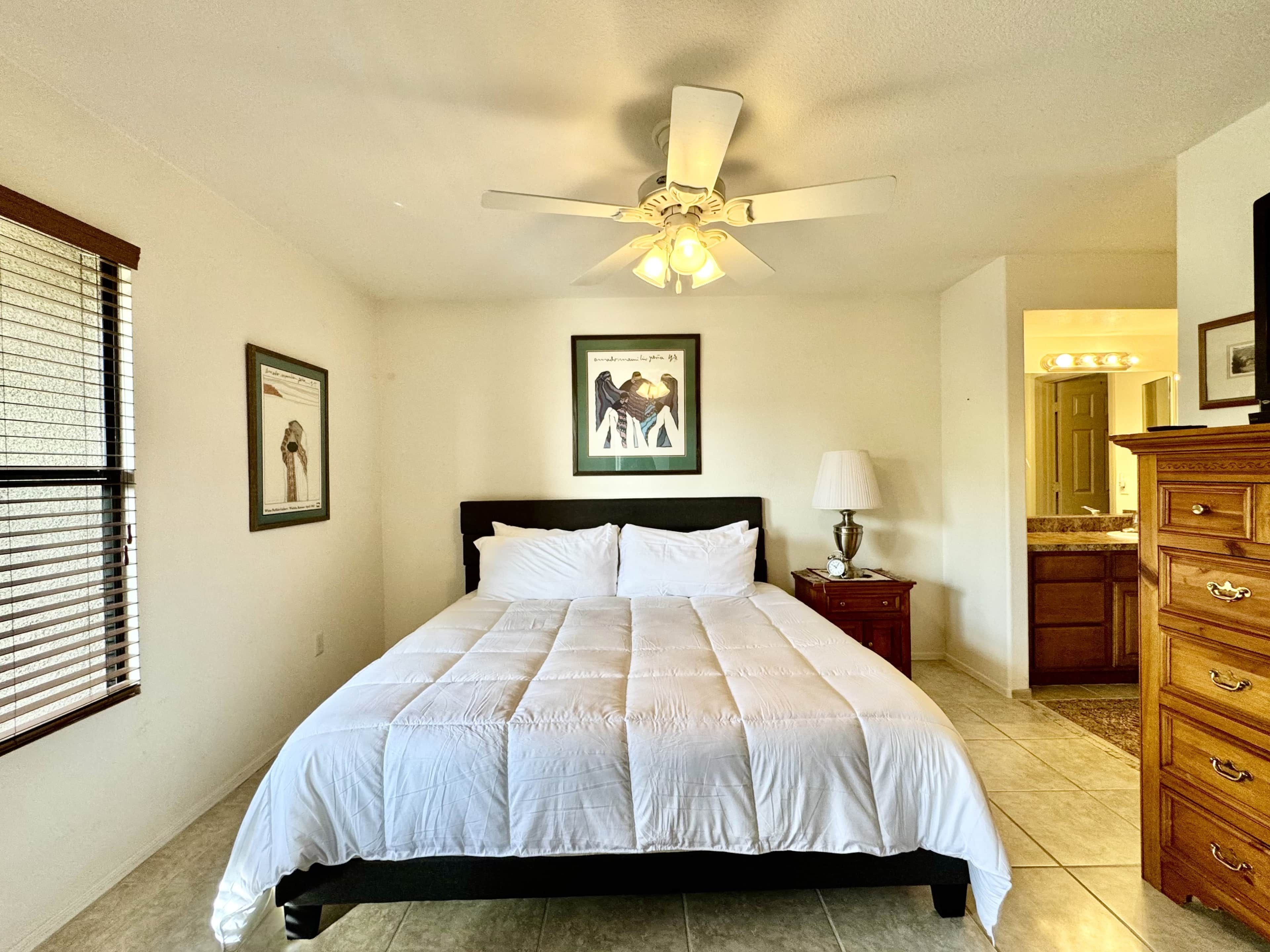 A bedroom with a bed, a ceiling fan, a nightstand with a lamp, a dresser, and framed pictures on the wall.