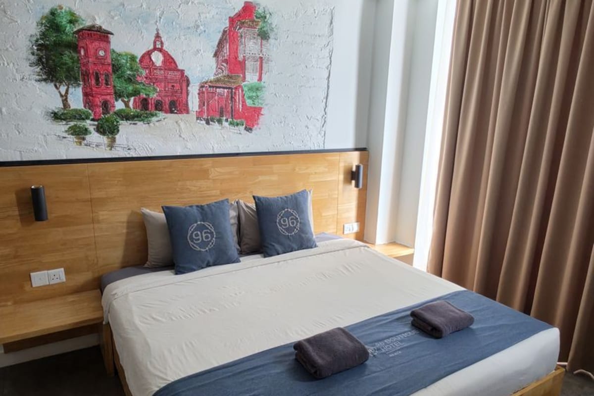 Habitació d'hotel amb un llit gran, un panell de fusta al capçal, un mural a la paret i una finestra amb cortines.