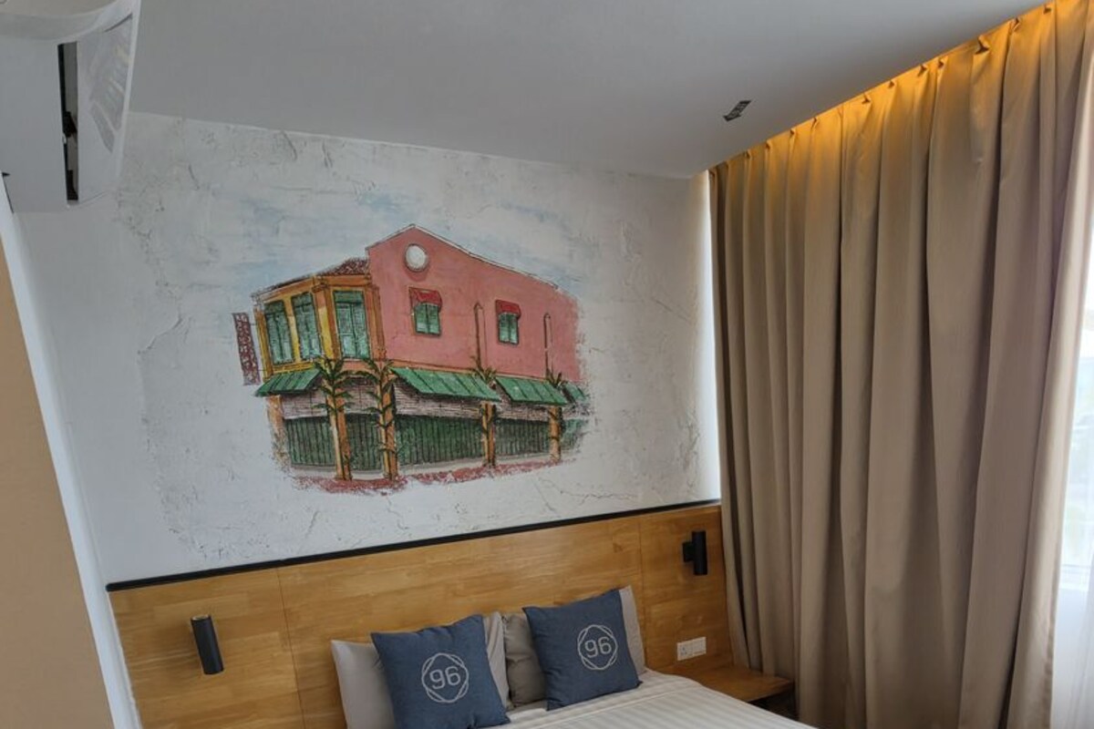 Ett hotellrum med en säng, en sänggavel, en väggmonterad luftkonditioneringsanläggning, ett fönster med gardiner och en väggdekoration som föreställer en byggnad.
